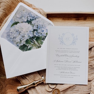 Puede incluir: Elegante juego de invitaciones de boda con una tarjeta blanca con un escudo floral y texto escrito. El sobre tiene un diseño floral en acuarela. También hay un sello de cera y sellos dorados.