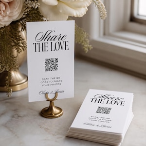 Könnte beinhalten: Eine weiße rechteckige Karte mit dem schwarzen Text Share the Love, einem QR-Code und den Namen Olivia & Lucas. Eine Karte steht aufrecht in einem kleinen goldfarbenen Metallständer, während ein Stapel identischer Karten daneben liegt.