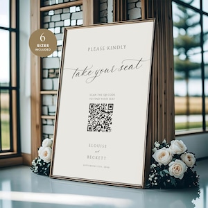 Puede incluir: Un letrero enmarcado para bodas con el texto "PLEASE KINDLY take your seat" en escritura cursiva. Incluye un código QR y los nombres "ELOUlSE and BECKETT". El letrero está enmarcado en dorado y decorado con rosas blancas y vegetación.