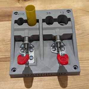 May include: A gray 3D-printed tool with two compartments, each secured by a metal latch with a red handle. A yellow cylinder is in one compartment. The tool is labeled "HARRIS 3D" and has markings for sizes 410, 28, and 12.