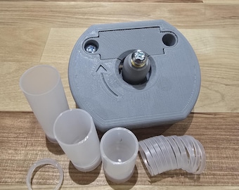 May include: A gray 3D-printed device with a central mechanism and an arrow indicator, accompanied by several translucent plastic cylinders, rings, and a coiled component. The items are arranged on a wooden surface.