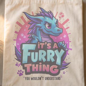 Può includere: Una borsa tote in tela bianca con un drago di cartone animato blu e viola e il testo "It's a Furry Thing You Wouldn't Understand".