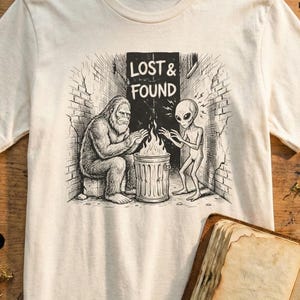 May include: Off-white t-shirt with a black and white illustration of a Bigfoot and an alien warming themselves by a fire. The text "LOST & FOUND" is above them. The shirt is on a wooden surface with a magnifying glass and other items.