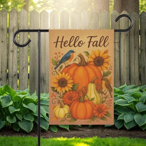 Puede incluir: Una bandera de jardín rectangular con temática otoñal. La bandera presenta las palabras "Hello Fall" sobre una ilustración de calabazas, girasoles y pájaros. La paleta de colores incluye tonos naranja, amarillo y marrón. La bandera se muestra en un soporte de metal negro.