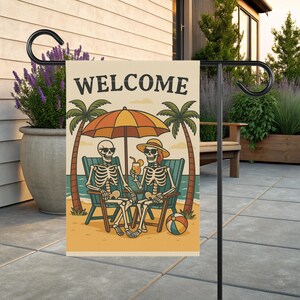 May include: A garden flag with the word "WELCOME" at the top. The flag features a cartoon illustration of two skeletons relaxing on beach chairs under an umbrella. Palm trees, the ocean, and a beach ball complete the scene.