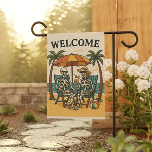Skeleton garden flag hanging on a yard pole, featuring two skeletons lounging under a colorful beach umbrella between palm trees on a sandy shore—ideal as a Summer Skeleton Flag, Spooky Garden Flag, Halloween Garden Flag, and Beach Skeleton Flag.
