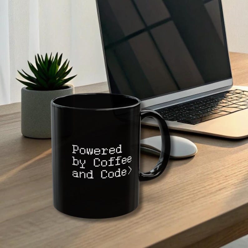 Black ceramic mug featuring pixel-style white text “Powered by Coffee and Code>” sitting on a warm wood desk beside a laptop, wireless mouse, and small succulent plant. Keywords: programmer gift, coder mug, developer coffee cup.