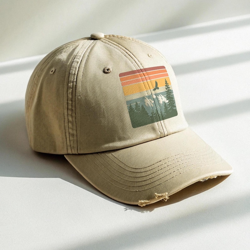 Light olive distressed dad hat resting in soft window light, showing a retro sunset-stripe mountain and forest patch graphic with a soaring eagle on the front panel.