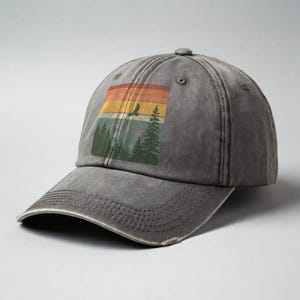 May include: A gray distressed baseball cap with a vintage-style graphic of a sunset, trees, and a bird flying over a mountain range.