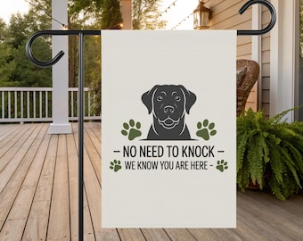 May include: A cream-colored garden flag featuring a black Labrador Retriever graphic and green paw prints. The flag has the text "- NO NEED TO KNOCK - WE KNOW YOU ARE HERE -" in black. The flag is displayed on a black metal stand.