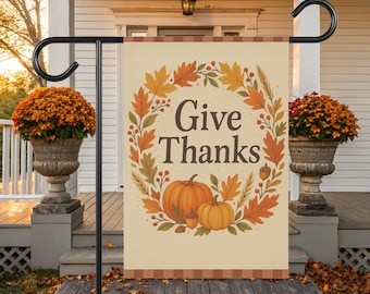 Könnte beinhalten: Ein rechteckiger Gartenflagge mit den Worten "Give Thanks" in brauner Schrift, umgeben von einem Kranz aus Herbstblättern, Kürbissen und Eicheln. Die Flagge ist cremefarben mit einem karierten Rand.