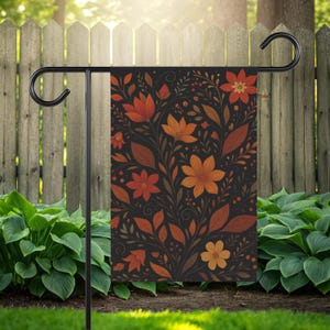 Könnte beinhalten: Eine schwarze Gartenflagge mit einem floralen Muster in Orange-, Rot- und Brauntönen. Das Design zeigt Blumen und Blätter in einem sich wiederholenden Muster. Die Flagge ist auf einem schwarzen Metallständer in einem Garten montiert.