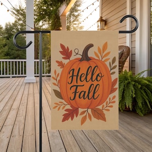 May include: A decorative outdoor flag featuring a large orange pumpkin with the words "Hello Fall" in elegant script. The pumpkin is surrounded by autumn leaves in shades of red, orange, and brown. The flag is displayed on a black metal stand.