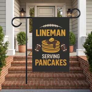 Puede incluir: Bandera de jardín negra con el texto "LINEMAN SERVING PANCAKES" en amarillo. El diseño incluye un balón de fútbol, una pila de panqueques con mantequilla y jarabe, y dos balones de fútbol. La bandera se muestra en un soporte de metal negro.