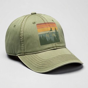Olive-green distressed dad hat on a neutral studio backdrop, featuring a retro square patch graphic of sunset stripes above pine trees, mountains, and a soaring eagle.