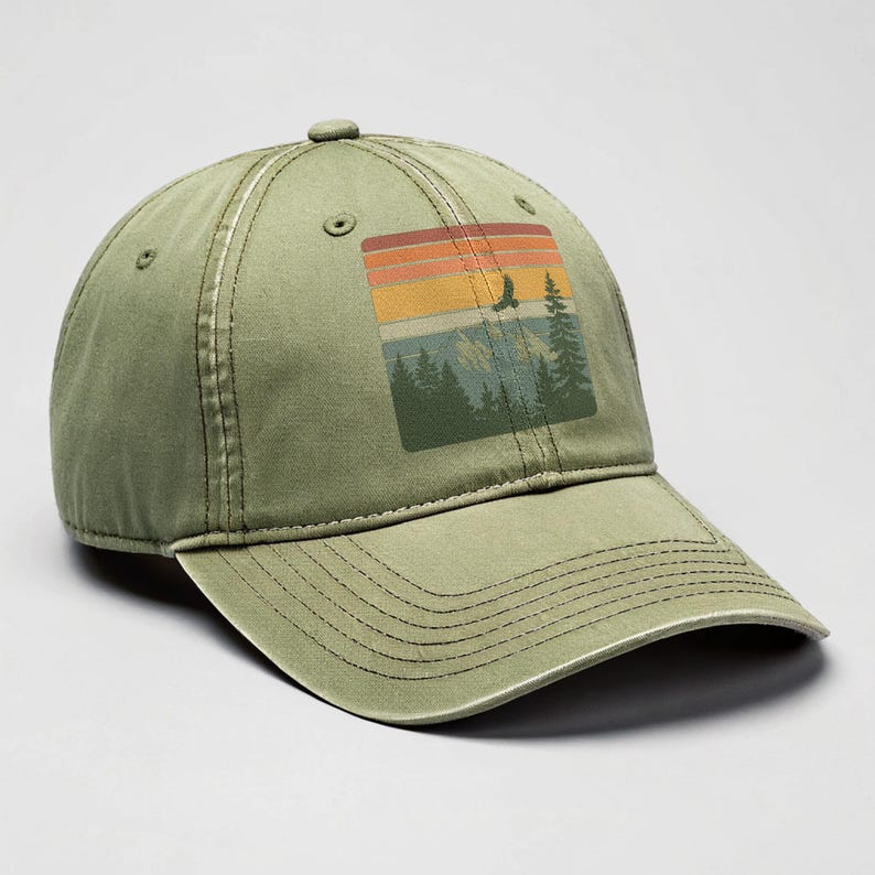 Olive-green distressed dad hat on a neutral studio backdrop, featuring a retro square patch graphic of sunset stripes above pine trees, mountains, and a soaring eagle.