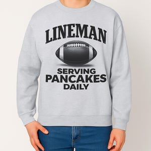 Op de afbeelding: Een lichtgrijs sweatshirt met de tekst "LINEMAN" boven een voetbalafbeelding en de woorden "SERVING PANCAKES DAILY" eronder. De voetbal is zwart en wit. De sweatshirt heeft een ronde hals.