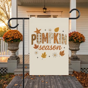 Puede incluir: Una bandera de jardín crema con las palabras "PUMPKIN season" en varios tipos de letra y gráficos otoñales. La bandera está montada en un soporte de metal negro. Dos grandes macetas llenas de flores naranjas flanquean la bandera.