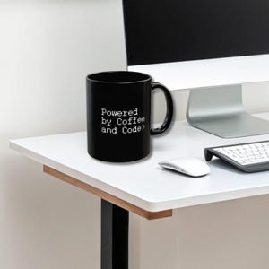 Black ceramic mug with pixel-style white text “Powered by Coffee and Code>” placed on a sleek white desk next to a wireless keyboard, mouse, and a modern computer monitor. Keywords: coder mug, programmer gift, developer coffee cup.