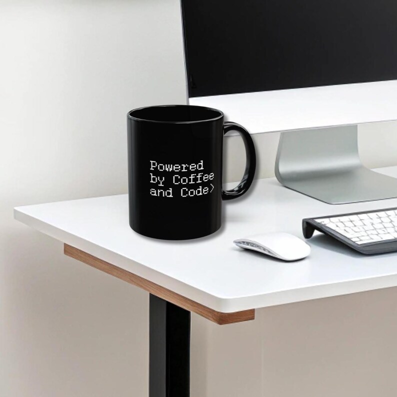 Black ceramic mug with pixel-style white text “Powered by Coffee and Code>” placed on a sleek white desk next to a wireless keyboard, mouse, and a modern computer monitor. Keywords: coder mug, programmer gift, developer coffee cup.