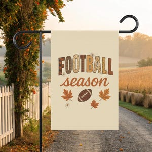 Puede incluir: Una bandera de jardín beige con las palabras "FOOTBALL season" en letras marrones y naranjas. La bandera presenta un gráfico de fútbol americano y hojas de otoño. La bandera está colgada de un soporte de metal negro.
