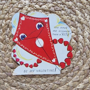 May include: Vintage Valentine's Day card featuring a red kite with a smiling face and heart-shaped accents. The card reads "You have me higher than a 'kite'" and "Be my Valentine!" on a woven background.