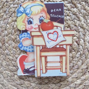 May include: Vintage Valentine's Day card featuring a cartoon girl with blonde hair and a blue bow. The card includes a desk, a red apple, and the text "Dear Teacher" and "Be My Valentine."
