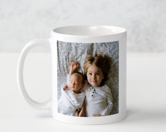 May include: White ceramic mug with a photo of two children, a baby and a toddler, lying on a patterned blanket. The toddler is smiling and looking at the camera. The baby is looking away from the camera.