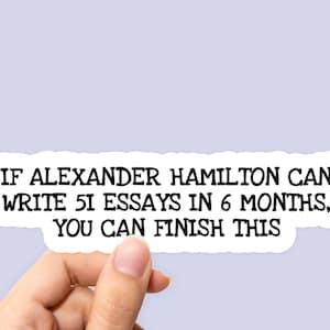 May include: A white sticker with black text that reads: "IF ALEXANDER HAMILTON CAN WRITE 51 ESSAYS IN 6 MONTHS, YOU CAN FINISH THIS." The sticker is held by a hand against a light purple background.