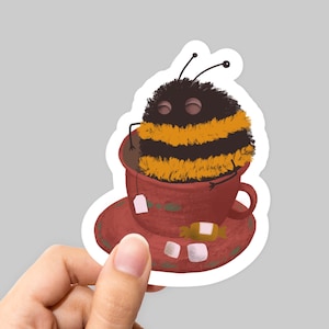 May include: A fuzzy yellow and black bumblebee sits in a red teacup with a tea bag and sugar cubes. The teacup is on a red saucer with a white rim.