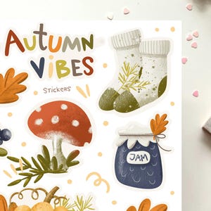 K&ouml;nnte beinhalten: Ein Set mit Herbst-Stickern, die einen Pilz, ein Glas Marmelade, ein Paar Socken und Bl&auml;tter zeigen. Die Sticker sind auf wei&szlig;em Hintergrund und im handgezeichneten Stil gehalten. Der Text "Autumn Vibes Stickers" befindet sich oben im Bild.