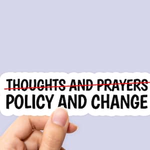 May include: White sticker with black text that reads "Thoughts and Prayers" with a red line through it and "Policy and Change" below. A hand is holding the sticker.