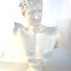 May include: White plaster bust sculpture of a man with short, curly hair. The bust is on a white pedestal.