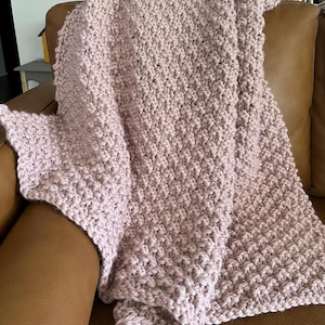 May include: A light mauve-colored, hand-knitted blanket draped over a brown leather couch. The blanket has a textured, woven appearance, with visible stitch patterns. The soft, cozy throw is folded, showcasing its thickness and warmth.