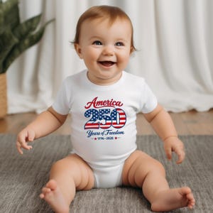 Może przedstawiać: Uśmiechnięte dziecko w białym body z napisem "America 250 Years of Freedom 1776-2026" w kolorze czerwonym i niebieskim. Dziecko siedzi na szarej macie.