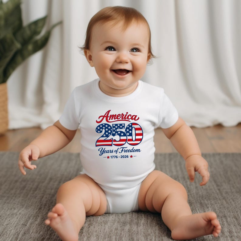 Może przedstawiać: Uśmiechnięte dziecko w białym body z napisem "America 250 Years of Freedom 1776-2026" w kolorze czerwonym i niebieskim. Dziecko siedzi na szarej macie.
