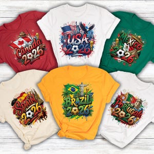 May include: A collection of short-sleeved t-shirts in various colors, each featuring a unique graphic design. The designs include country flags, soccer balls, and the year "2026". The shirts are in red, white, green, yellow, and beige.