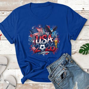 May include: A royal blue t-shirt featuring a graphic design with the text "USA 2026". The design includes an American flag, an eagle, a soccer ball, and the Statue of Liberty. The shirt is paired with denim shorts.