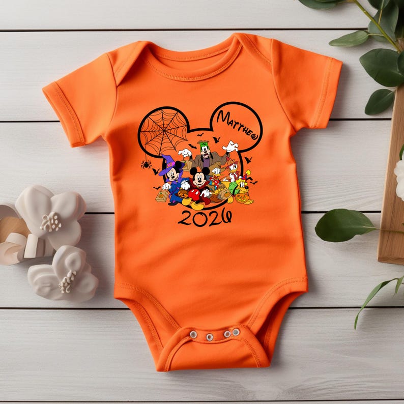 May include: An orange short-sleeved baby bodysuit featuring a black graphic of Mickey Mouse and friends in Halloween costumes inside a Mickey-shaped outline. The design includes a spider web, bats, the name Matthew, and the year 2026 in black text.