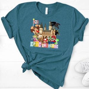 Puede incluir: Camiseta de color turquesa con un dise&ntilde;o gr&aacute;fico colorido. El dise&ntilde;o incluye personajes de dibujos animados, un castillo y el texto "EPIC UNIVERSE". Una peque&ntilde;a bandera indica "2026".