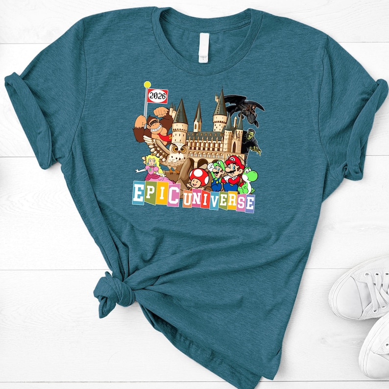 Puede incluir: Camiseta de color turquesa con un dise&ntilde;o gr&aacute;fico colorido. El dise&ntilde;o incluye personajes de dibujos animados, un castillo y el texto "EPIC UNIVERSE". Una peque&ntilde;a bandera indica "2026".