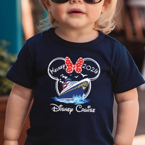 May include: Navy blue t-shirt featuring a Disney Cruise design. The design includes a cruise ship, the text "Disney Cruise", the name "Kelsey", the year "2020", and a red polka dot bow. The shirt is designed for children.