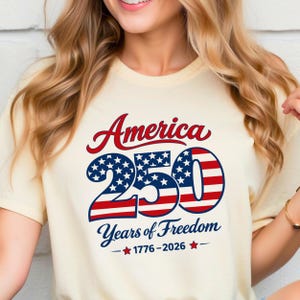 Może przedstawiać: Kremowa koszulka z napisem "America 250 Years of Freedom" w kolorze czerwonym i niebieskim. Liczba 250 jest zaprojektowana z gwiazdami i paskami, upamiętniając lata 1776-2026.