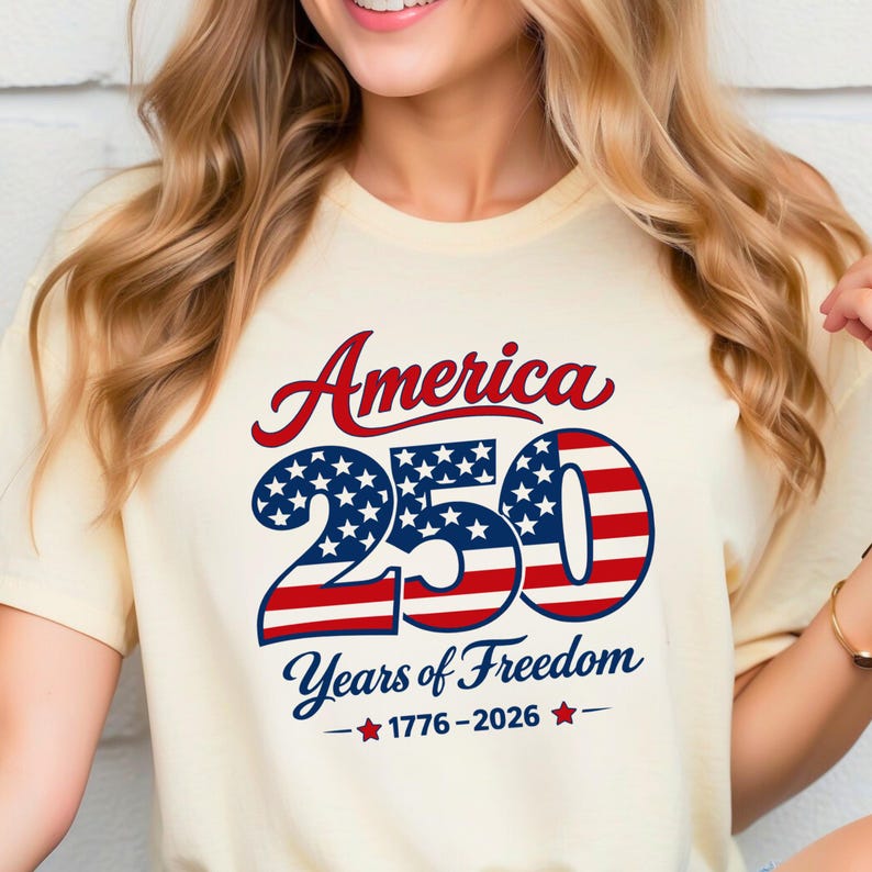 Może przedstawiać: Kremowa koszulka z napisem "America 250 Years of Freedom" w kolorze czerwonym i niebieskim. Liczba 250 jest zaprojektowana z gwiazdami i paskami, upamiętniając lata 1776-2026.