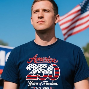 Może przedstawiać: Granatowa koszulka z napisem "America 250 Years of Freedom" w kolorze czerwonym, białym i niebieskim. Projekt zawiera gwiazdy i paski, z latami 1776-2026 poniżej. W tle widoczna jest amerykańska flaga.