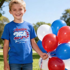 Może przedstawiać: Dziecko w niebieskiej koszulce z napisem "America 250 Years of Freedom 1776-2026" w kolorach czerwonym, białym i niebieskim. Trzyma bukiet czerwonych, białych i niebieskich balon&oacute;w, świętując patriotyczny motyw.