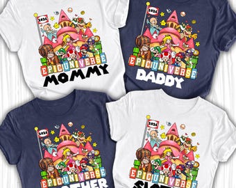 Op de afbeelding: Vier witte en marineblauwe t-shirts met een kleurrijke cartoonafbeelding. Elk shirt toont een kasteelscène met personages en de tekst "EPIC UNIVERSE" en de woorden "MOMMY", "DADDY", "BROTHER" en "SISTER".