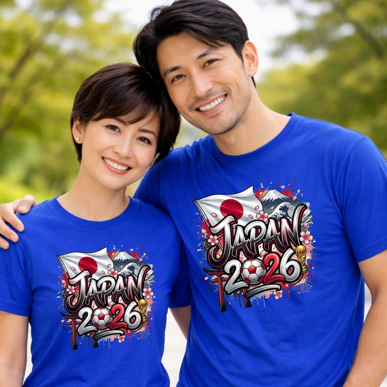 May include: Two people wearing blue t-shirts with a graphic design. The design includes the Japanese flag, Mount Fuji, cherry blossoms, and the text "JAPAN 2026" with a soccer ball. The shirts are short-sleeved.