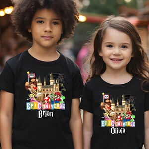 Puede incluir: Dos ni&ntilde;os vistiendo camisetas negras con un gr&aacute;fico colorido de "Epic Universe". El gr&aacute;fico incluye un castillo, varios personajes de dibujos animados y los nombres "Brian" y "Olivia". Tambi&eacute;n es visible un peque&ntilde;o "2026".