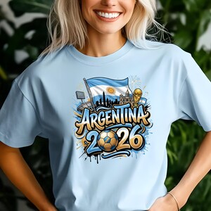 May include: Light blue t-shirt featuring a graphic design with the word "Argentina" and the number "2026". The design includes a soccer ball, the Argentinian flag, and a silhouette of a city skyline. A gold trophy is also visible.